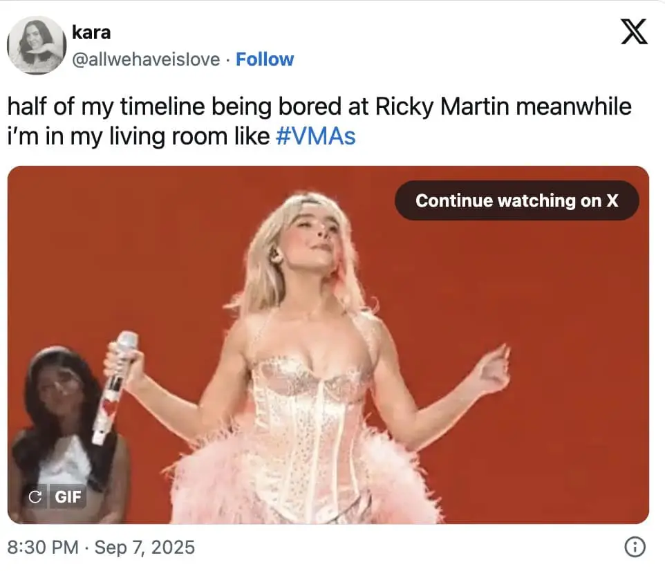 Pop performer in a pink corset with feather bustle poses on a red-lit stage, mic in hand