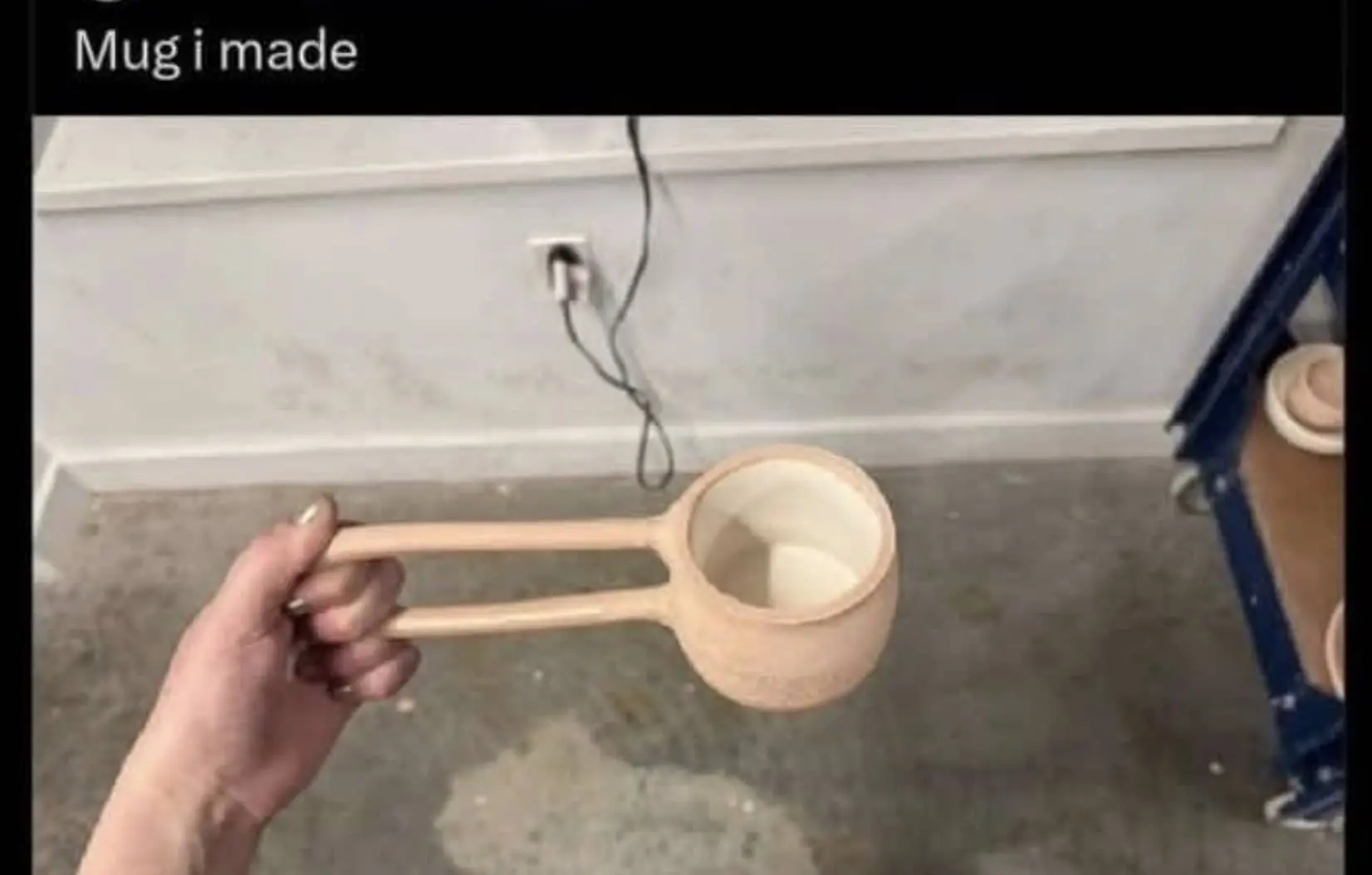 A handmade ceramic mug with two long, thin horizontal parallel handles being held against a workshop background.