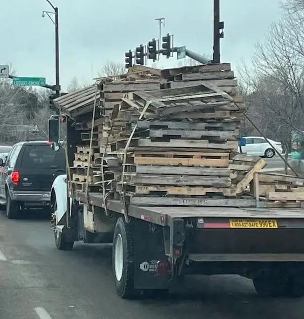A high-stakes funny osha violation picture of a flatbed truck transporting a precarious mountain of wooden pallets stacked three times higher than the cab, held together by a single, desperate tension strap that is doing the absolute most to prevent a total road hazard.