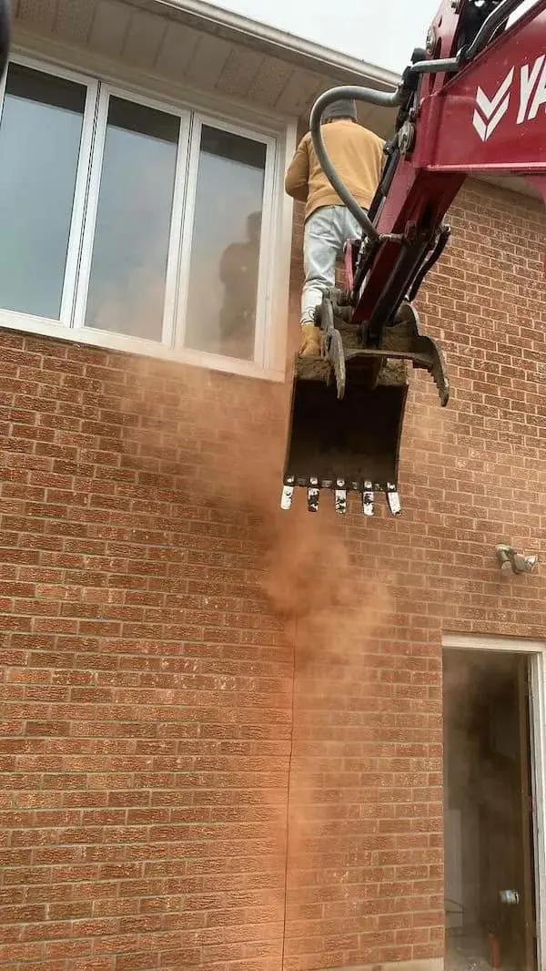 A funny Osha violation picture of a construction worker using an excavator bucket as a makeshift aerial lift to reach a second-story window, perfectly capturing the spirit of improvised workplace hazards.