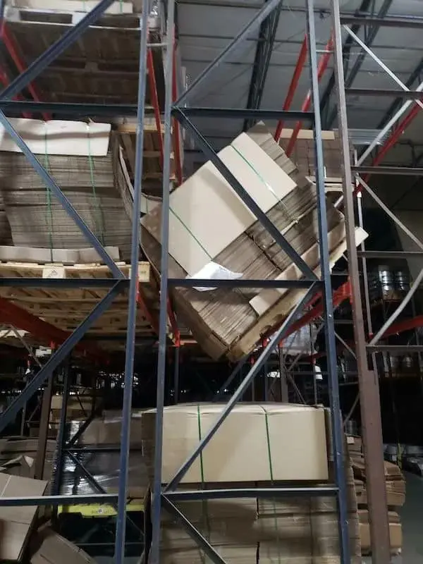 A warehouse nightmare funny osha violation picture showing a massive, multi-ton bale of cardboard wedged diagonally inside a racking system, held up by sheer friction and the structural integrity of a single bent metal beam.