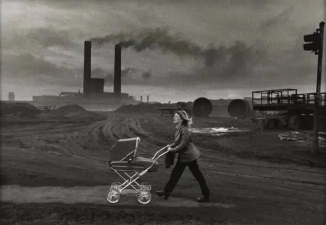Black and white photo of woman pushing pram past smoking industrial chimneys on barren wasteland