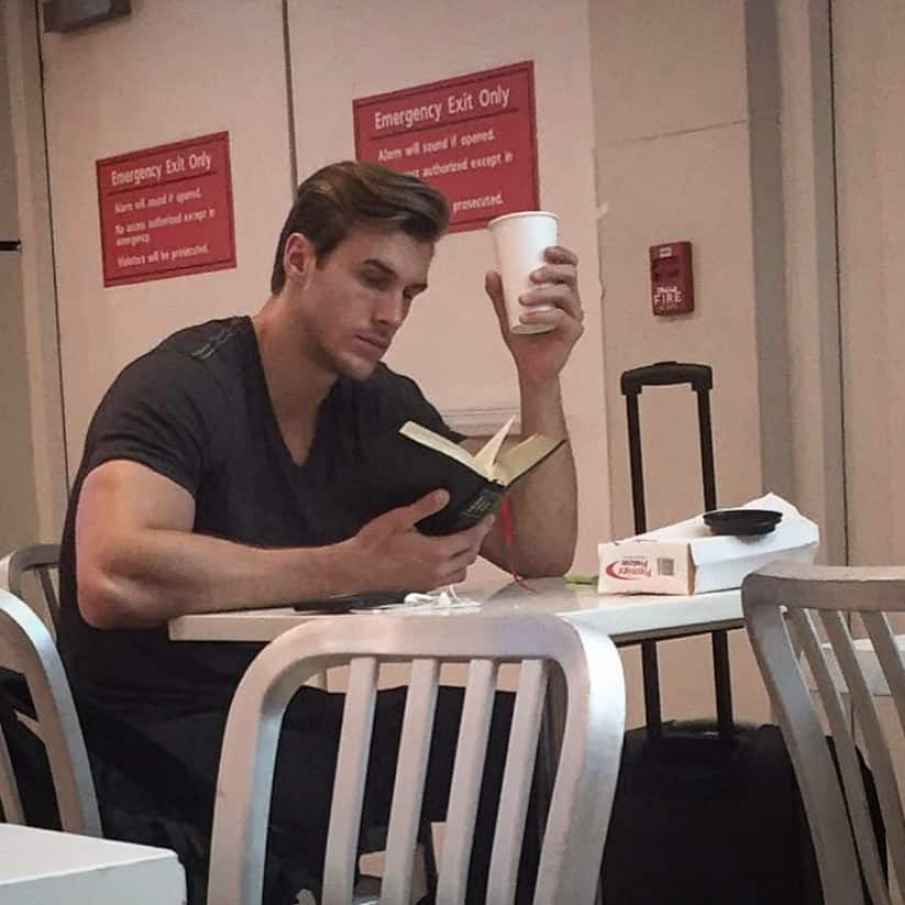 Man reading a book alone at airport table with suitcase beside him