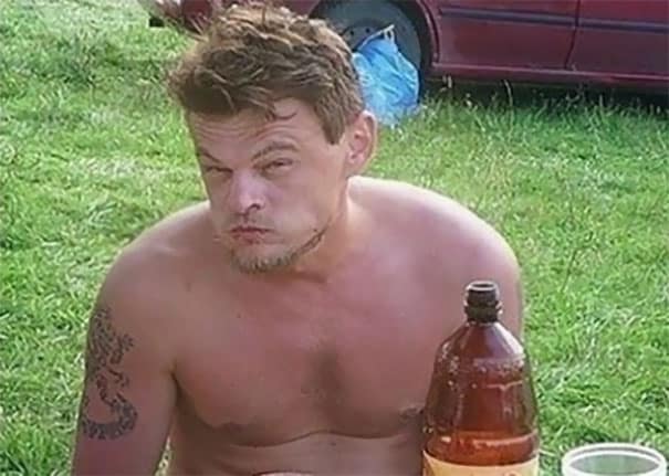 Shirtless man with a tattoo making a puffy face while sitting outdoors with a drink.