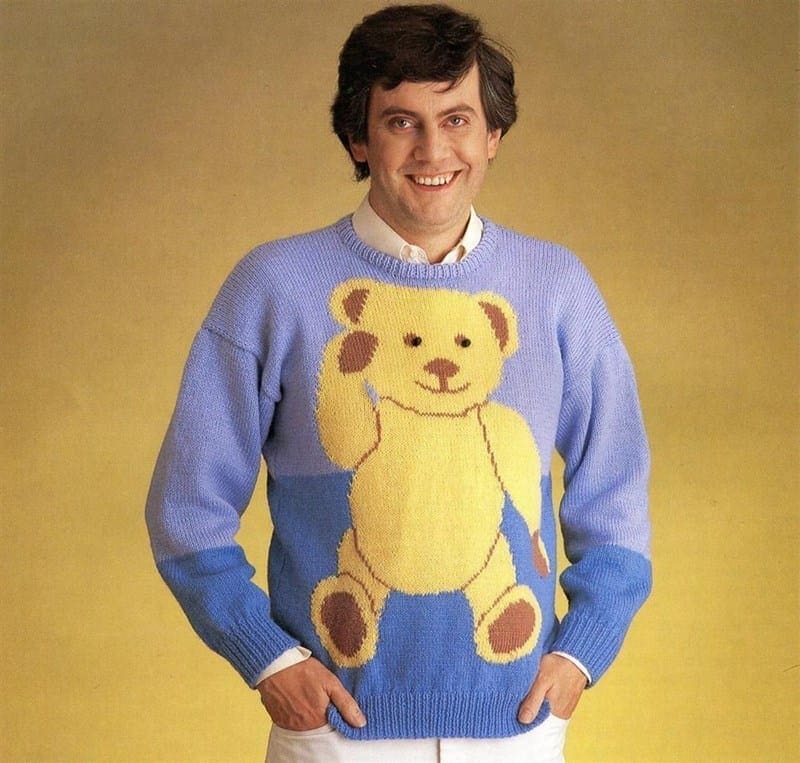 A man in a purple and blue sweater with a huge teddy bear graphic, posing with hands in his pockets.