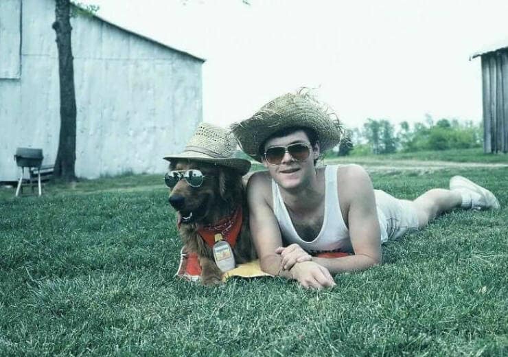 Man and dog wearing matching sunglasses and straw hats, lying in the grass