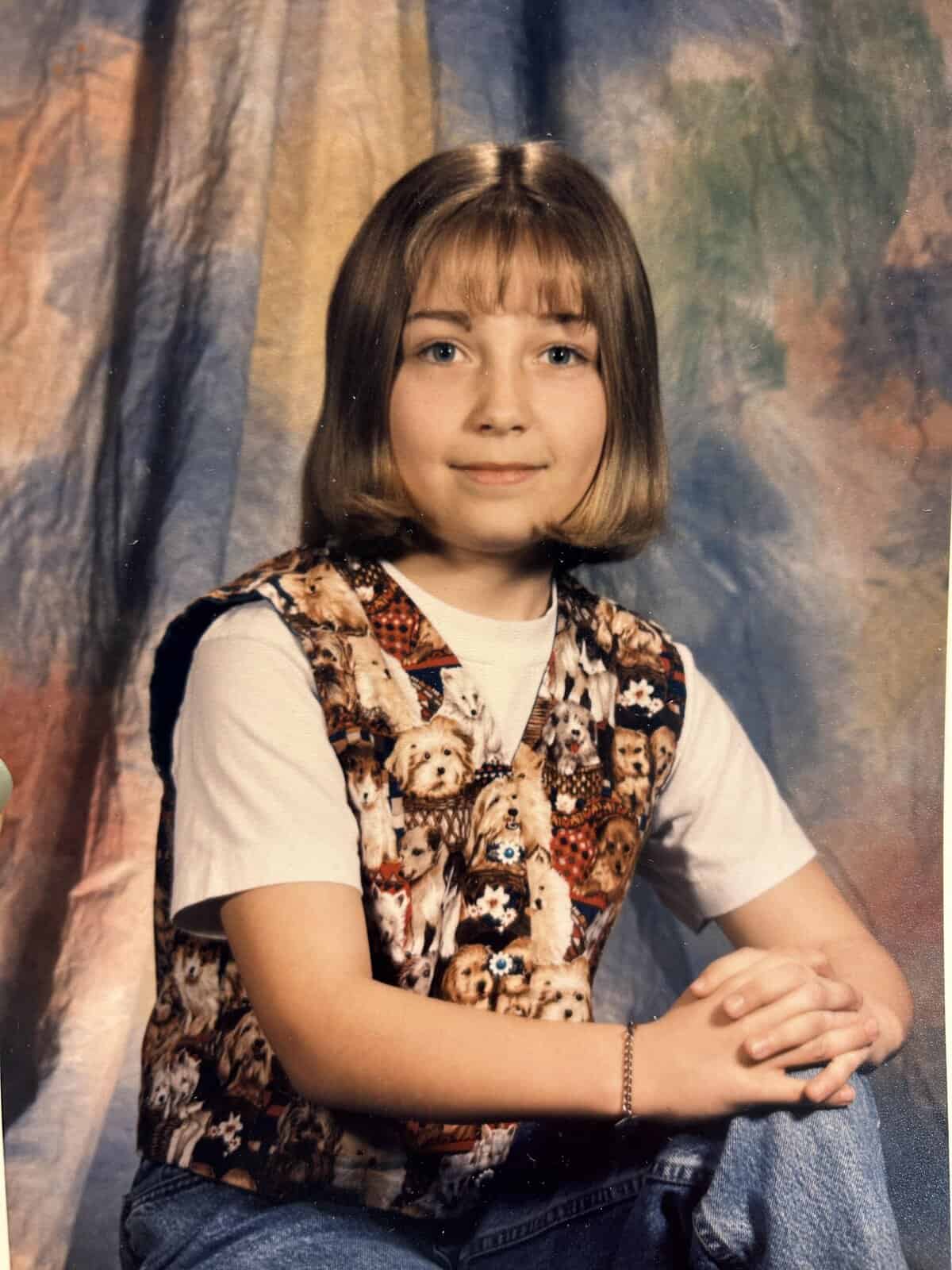 School portrait of a kid with a bob haircut wearing a vest covered in dog faces, hands clasped, mottled backdrop.