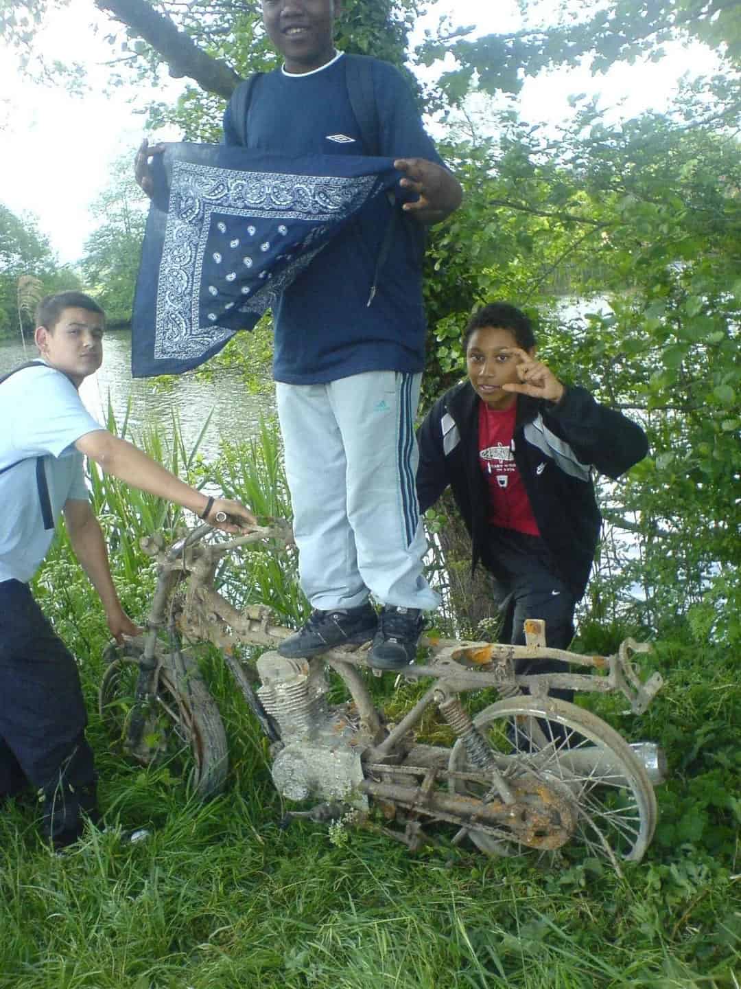 Three kids by a river posing with a rusty motorcycle frame; one stands on it while holding a blue bandana.