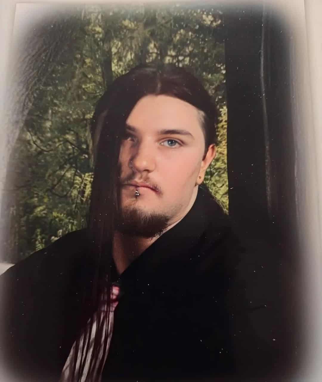 Moody studio portrait of a teen with side-swept emo hair, lip piercing, and goatee against a forest backdrop.
