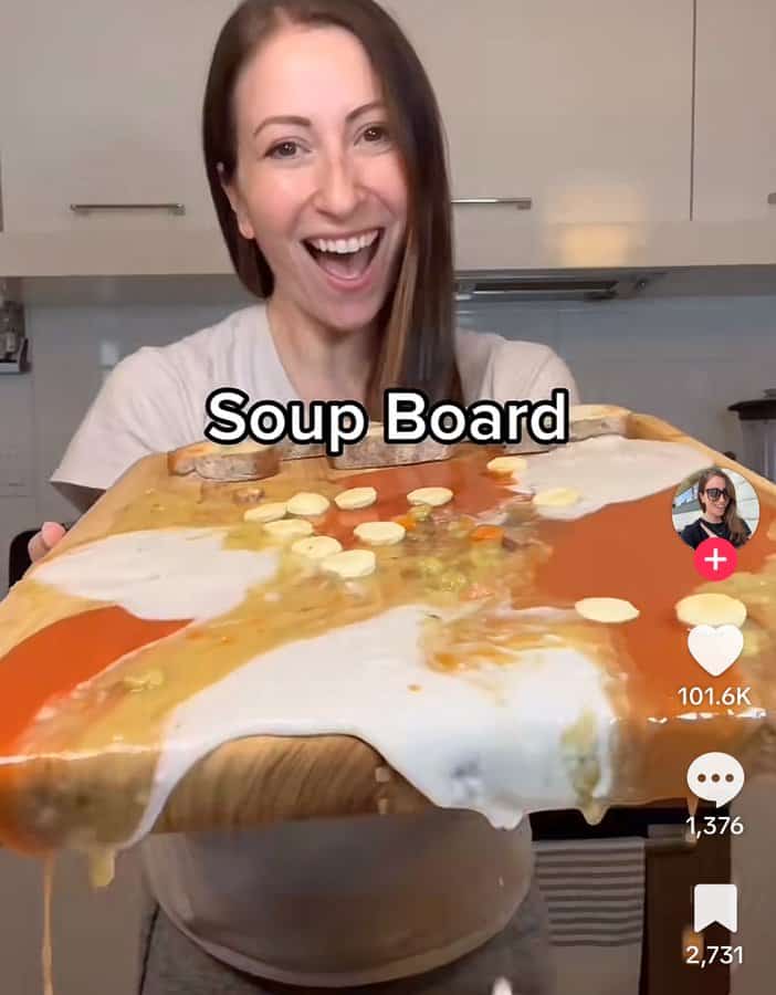Cursed screenshot of a woman proudly holding a messy “soup board” with different soups spilling off a cutting board.