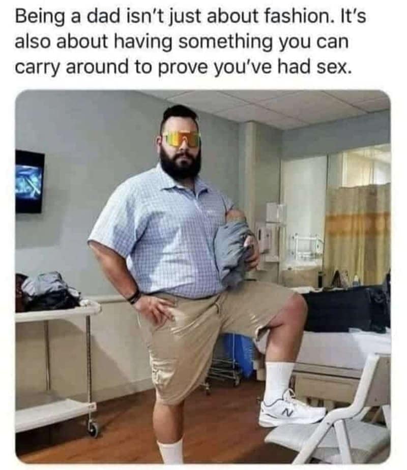 New dad in cargo shorts and shades posing with a baby carrier in a hospital, striking a confident stance.