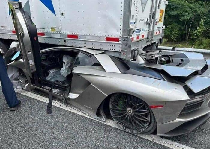 A silver Lamborghini Aventador supercar wedged and crushed underneath the back of a semi-truck.