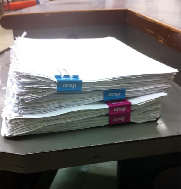 Stacks of assignments clipped with binder clips labeled “crap,” lined up on a desk.