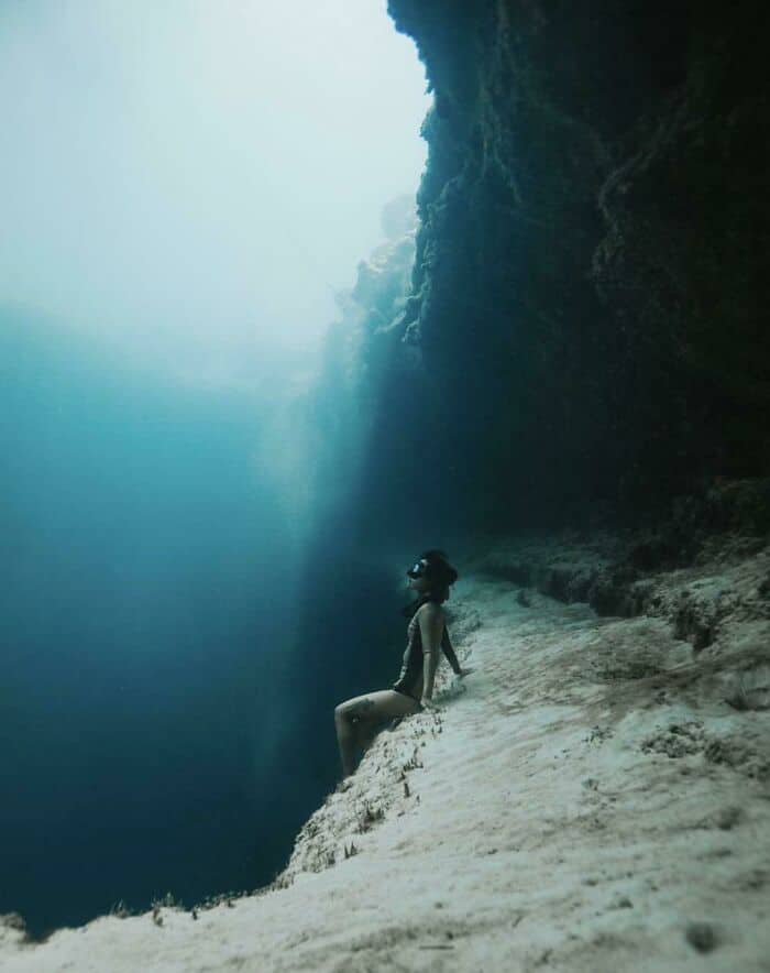 A woman sits on a sandy underwater cliff, looking down into a vast, dark ocean abyss.