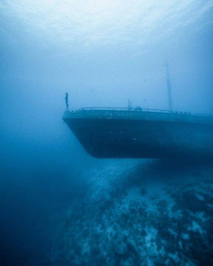 A ghostly shipwreck emerges from the murky blue depths with a lone diver on its bow.