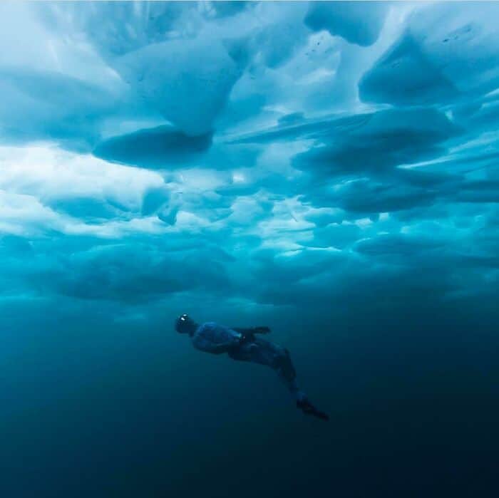 A free diver swims in the deep blue water underneath a vast, solid ceiling of cracked ice.
