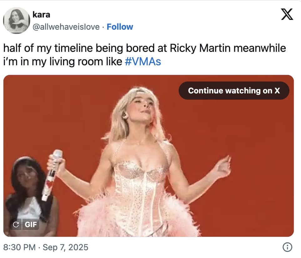 Pop performer in a pink corset with feather bustle poses on a red-lit stage, mic in hand