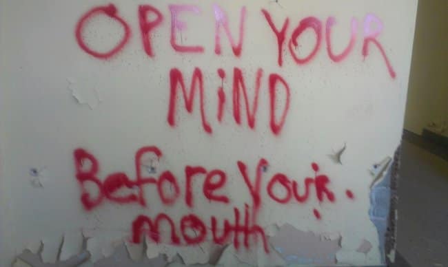 A piece of red spray paint graffiti on a dirty and decaying wall that offers the simple and profound piece of advice, "Open your mind before your mouth."