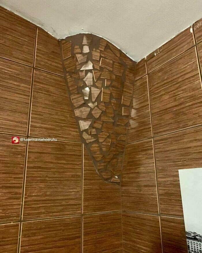 A terrible tile repair job in a brown, wood-grain tiled shower corner that uses a chaotic mosaic of broken tile pieces and dark grout.
