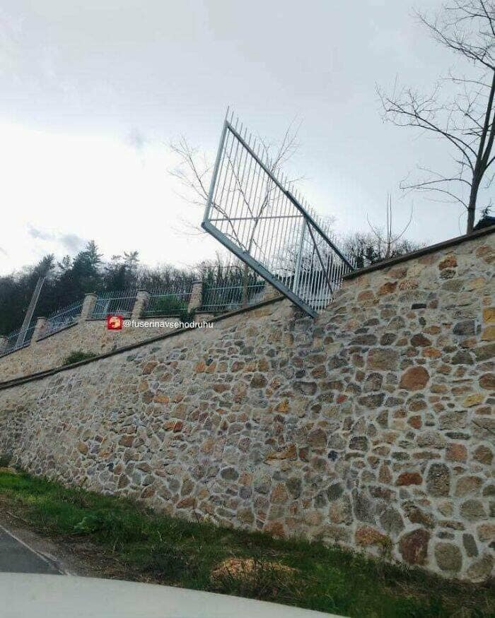 A section of a metal fence on top of a stone wall that has been bizarrely and incorrectly installed at a steep diagonal angle.