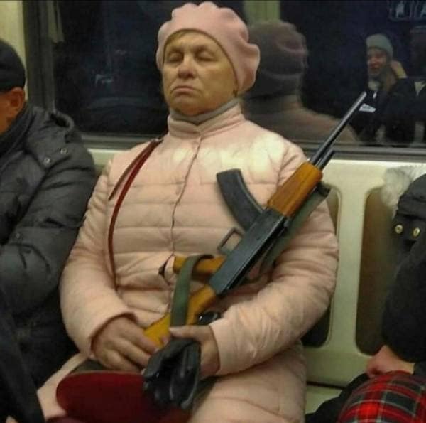 A very shocking and confusing photograph of a woman who is wearing a pink quilted jacket and is sleeping on the subway while calmly holding a large assault rifle in her lap.