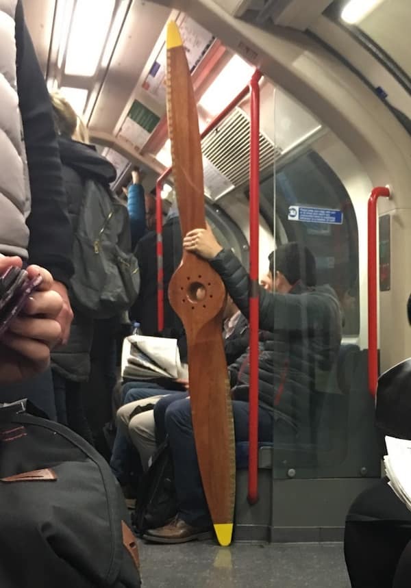A funny and bizarre photo of a man who is casually holding a giant, wooden, old-fashioned airplane propeller while he is sitting on a very crowded subway train in London.