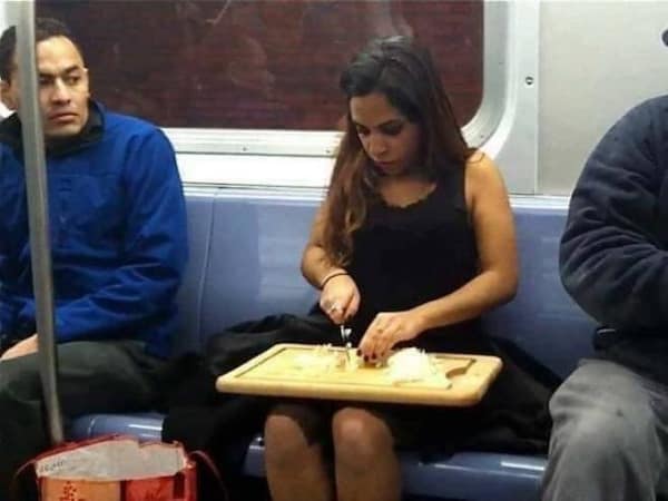A photograph of a woman who is using a large kitchen knife and a wooden cutting board to chop up some vegetables on her lap while she is riding the subway during her commute.