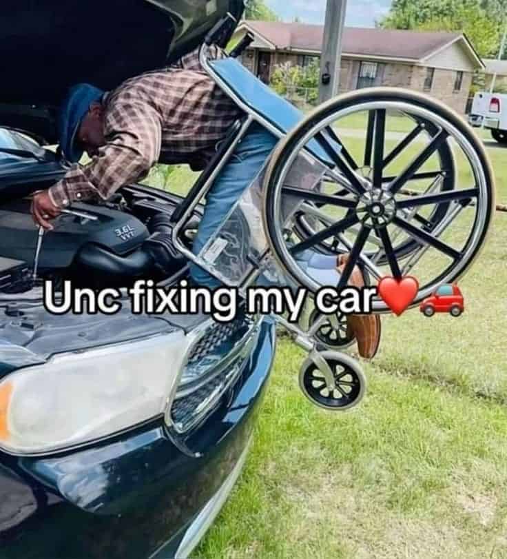 An impressive and wholesome photo of a man in a wheelchair leaning far over into a car's engine bay to fix it.