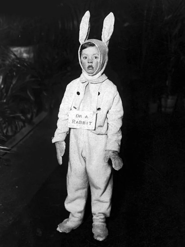 A funny, old, black-and-white photograph of a young child who is in a rabbit costume and is wearing a helpful handmade sign that says, "I'M A RABBIT," just in case people were confused.