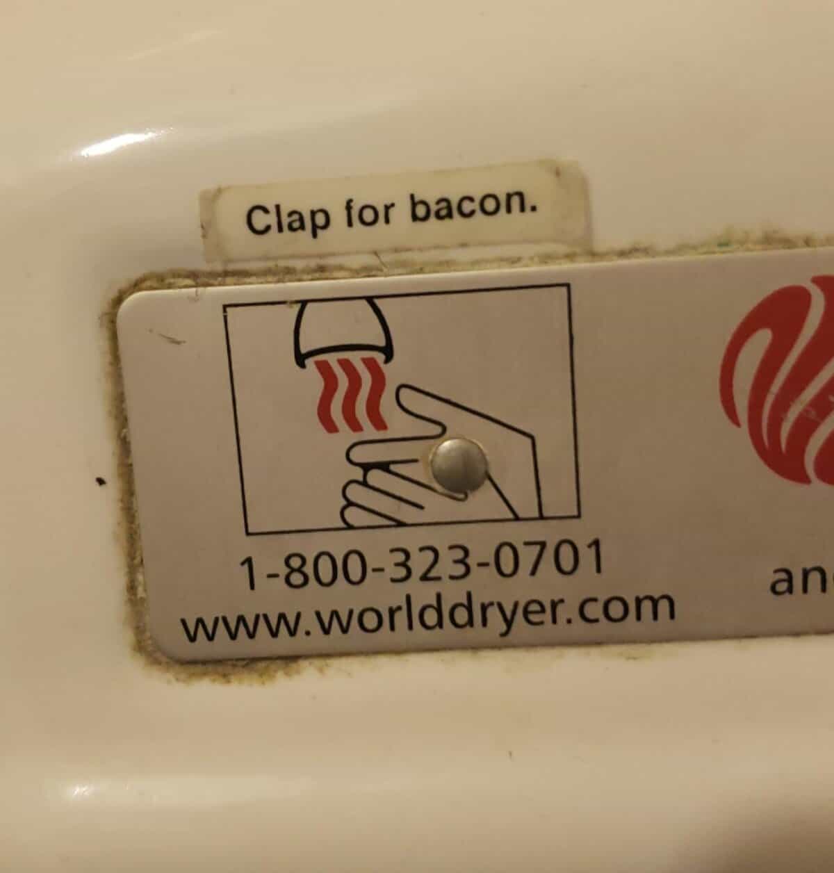 A funny piece of wholesome vandalism where a sticker saying "Clap for bacon" is placed on a public hand dryer.