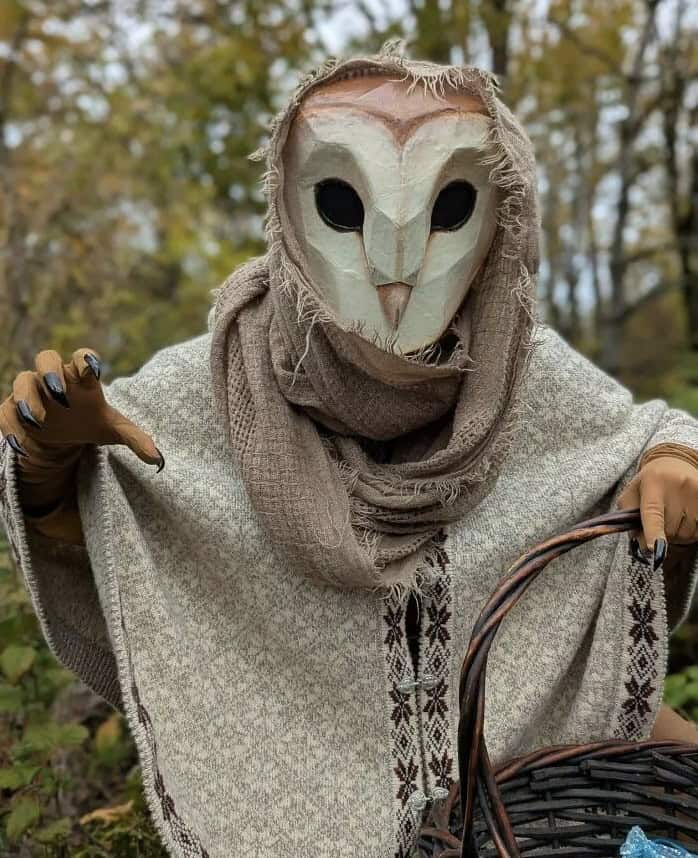 A person wearing a creepy barn owl mask, shawl, and gloves, holding a wicker basket.