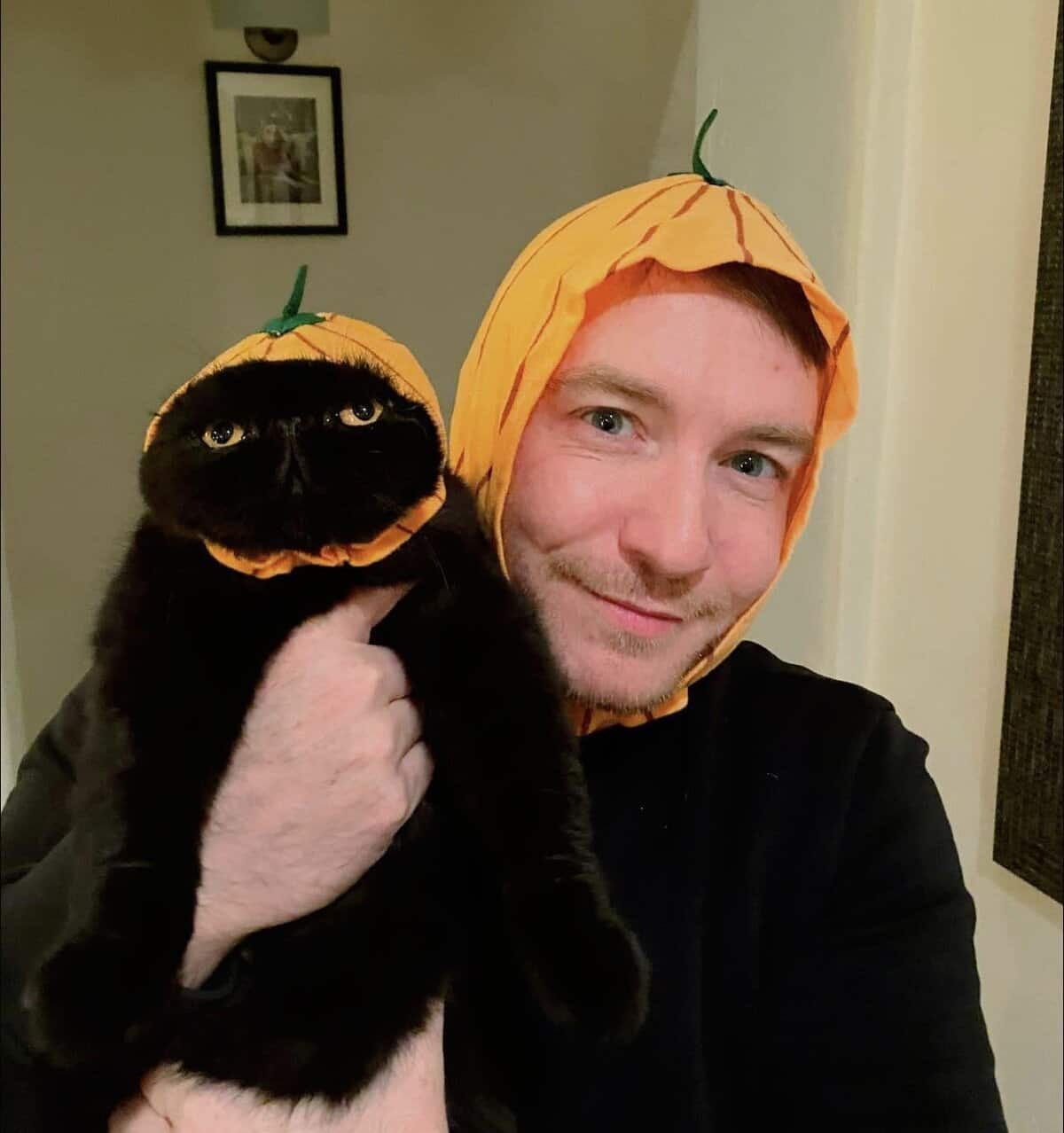 A man holding his black cat while they both wear matching orange pumpkin hoods.