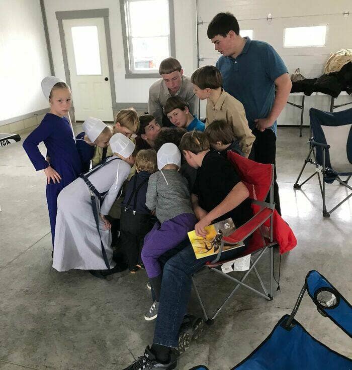 A group of curious Amish children gather around one boy sitting and reading a comic book.