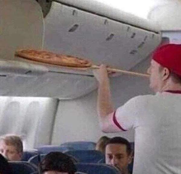 A person using a pizza peel to carefully place a whole pizza into an airplane's overhead bin compartment.