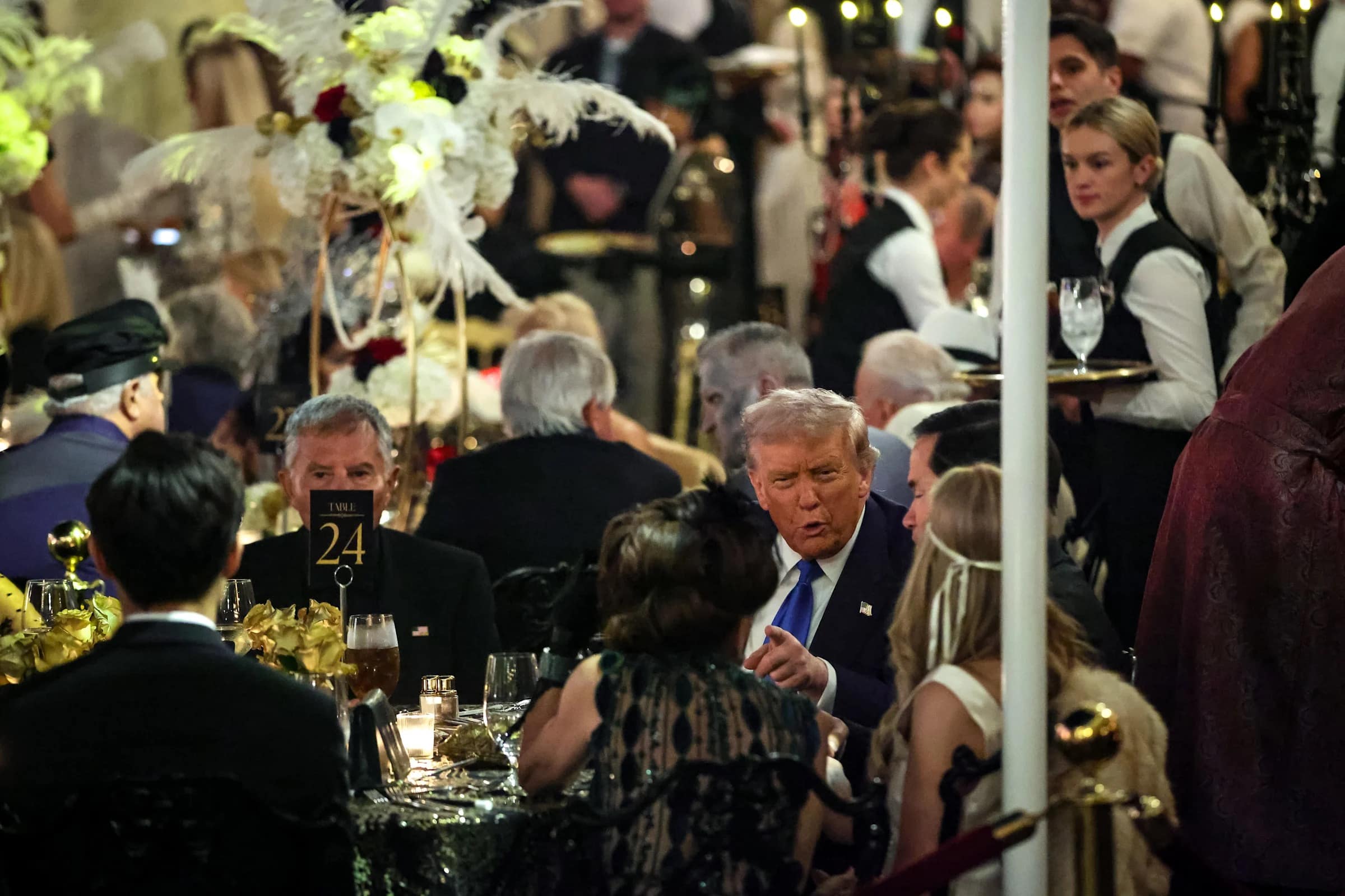 A photo of Donald Trump at his table during the Mar-a-Lago Great Gatsby party, talking to guests.