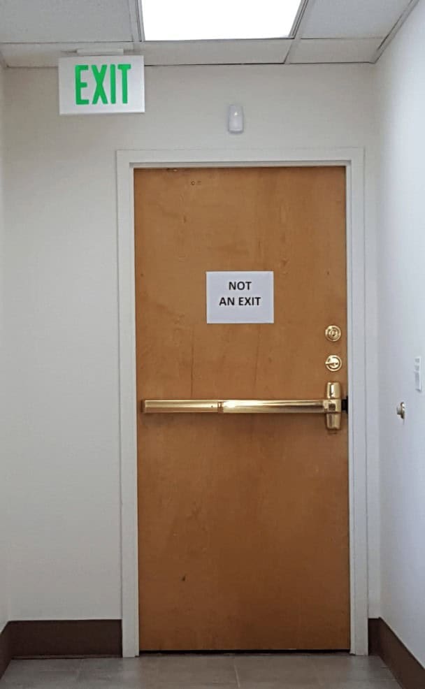 A hilarious and funny design fail showing a door with a glowing "EXIT" sign above it and a paper "NOT AN EXIT" sign on it.