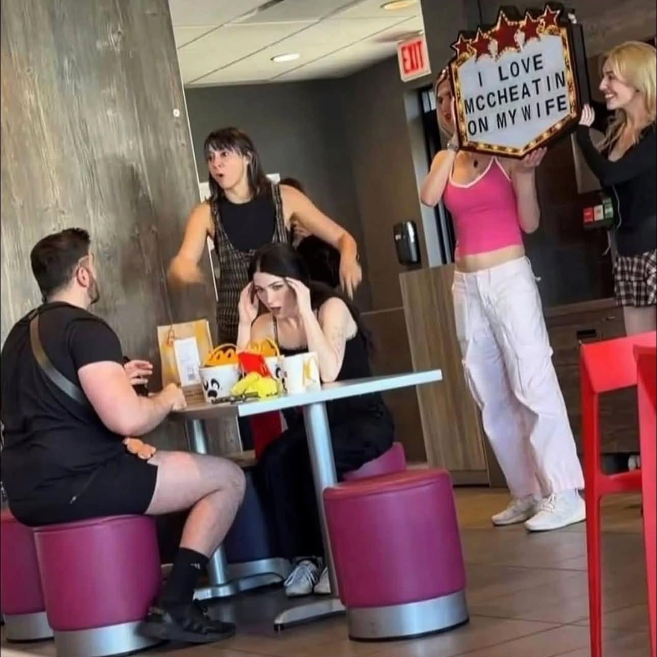 Funny candid photo of women confronting a man at McDonald's with a "I Love McCheatin" sign.