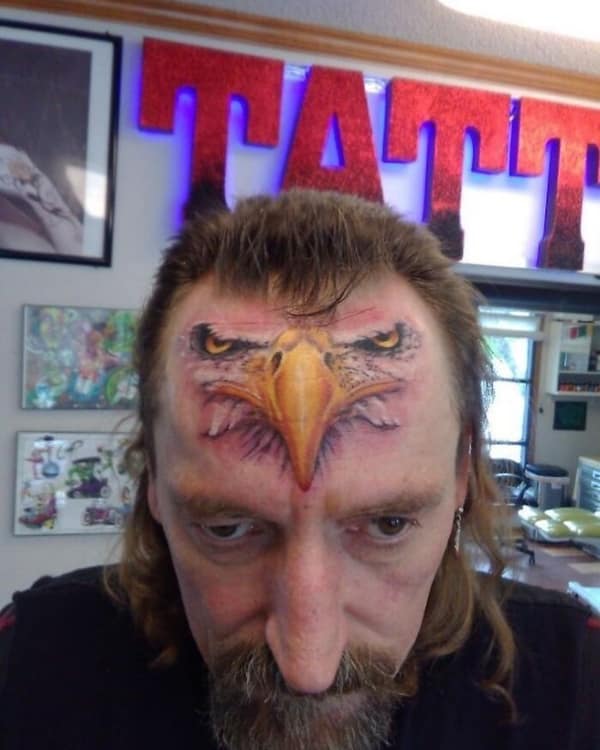 Realistic eagle face tattoo covering a man's entire forehead and nose bridge.