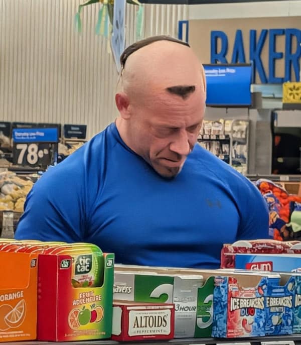 A muscular, bald man in a Walmart with a few tiny, random patches of hair left on his head.