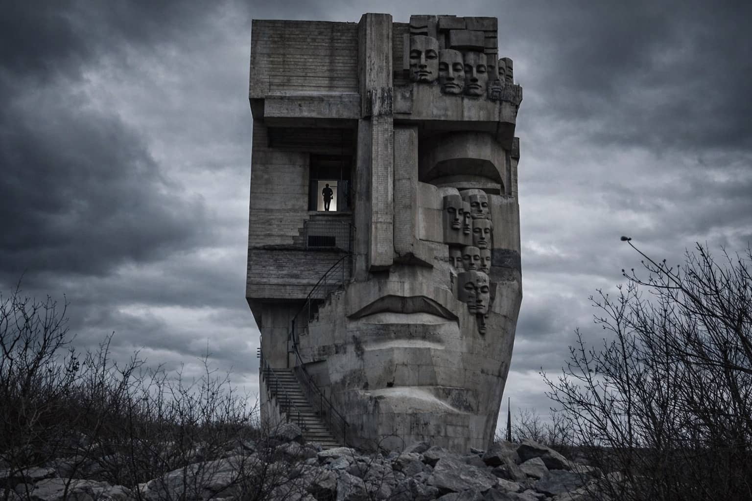 Brutalist evil architecture concrete building shaped like a giant human face with multiple smaller masks.