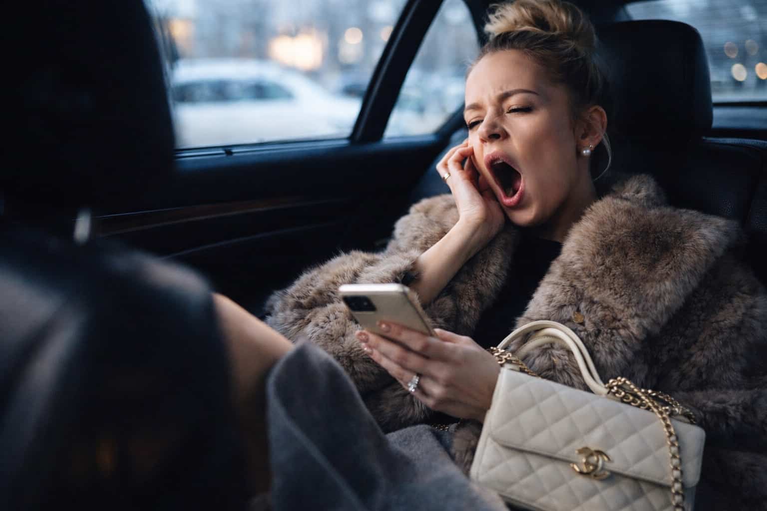 Woman in a fur coat yawning while looking at her phone in the back of a car.