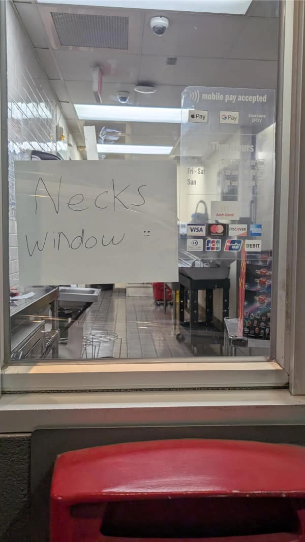A simple but effective english fail showing a handwritten sign taped to a fast-food drive-thru glass that incorrectly directs customers to the "Necks Window."