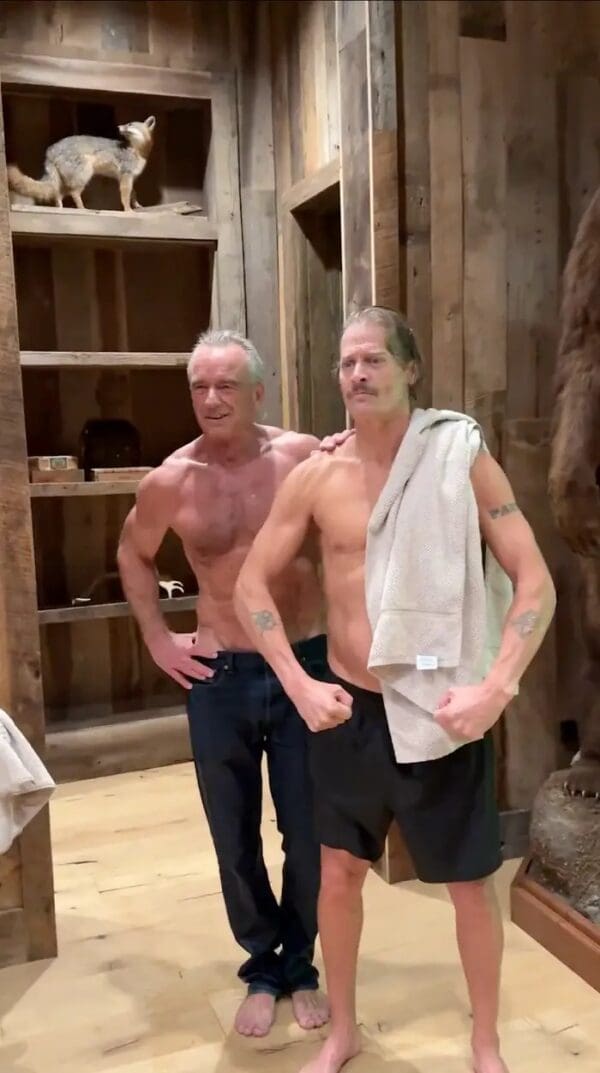 A bizarre RFK JR and Kid Rock workout photo showing a shirtless Robert F. Kennedy Jr. wearing blue jeans alongside a shirtless Kid Rock flexing in a rustic wood-paneled room surrounded by taxidermy animals.