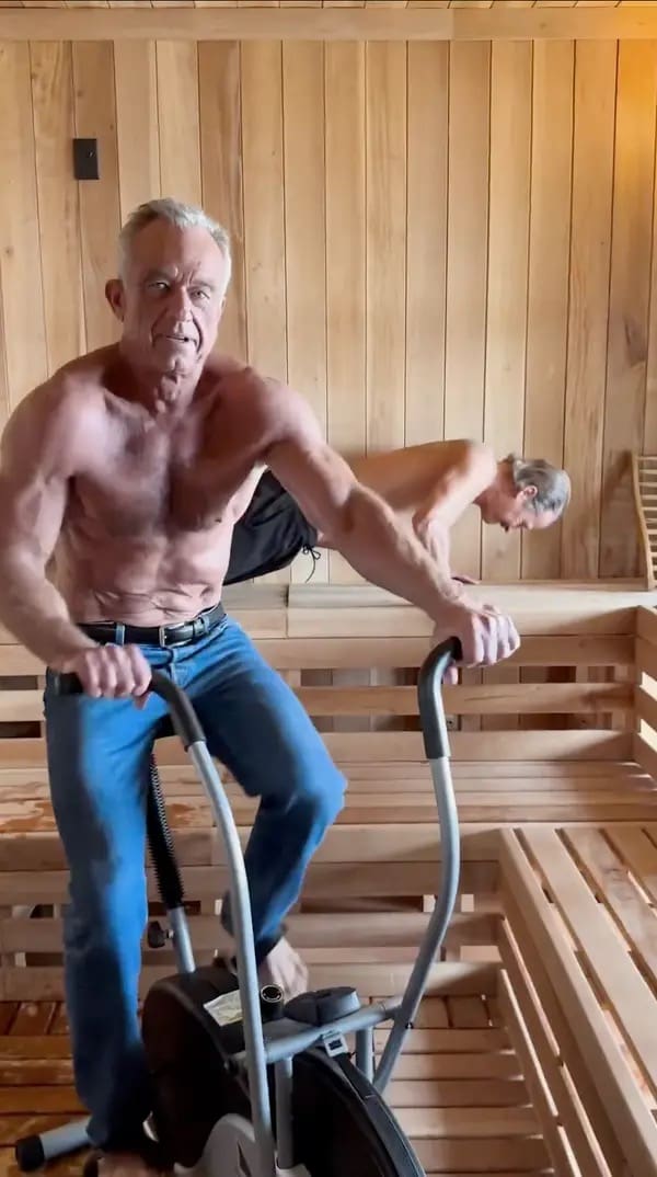 An absurd RFK Jr sauna photo capturing a sweating, shirtless RFK Jr. vigorously riding a stationary exercise bike inside a wooden sauna while inexplicably wearing blue jeans and a black leather belt.