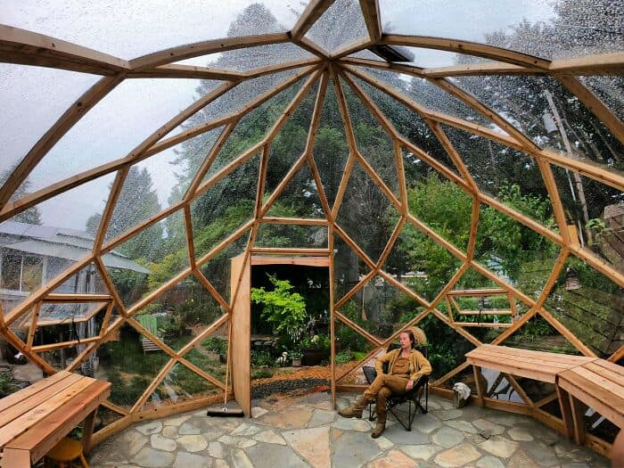 Geodesic dome greenhouse built with a complex timber frame and clear panels. A woman sits inside this backyard retreat.