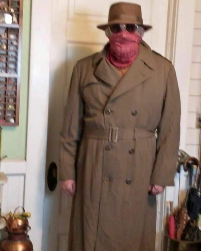Photo of a person wearing a trench coat, hat, sunglasses, and a red face covering indoors.