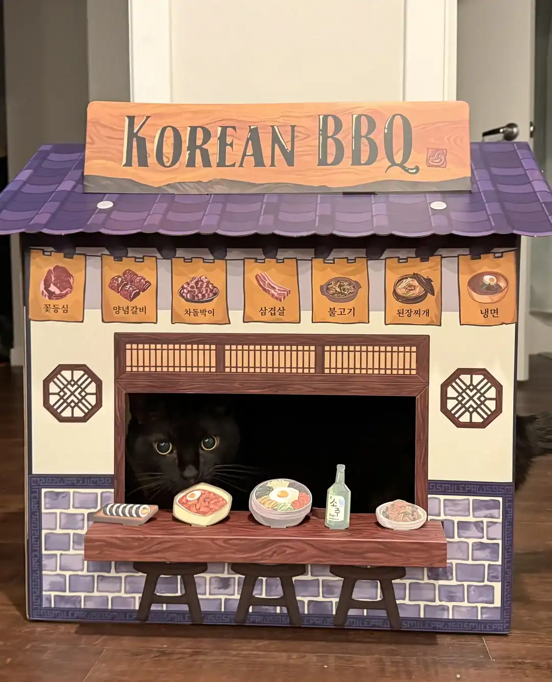 Black cat looking out from a detailed cardboard Korean BBQ restaurant stand with food illustrations.