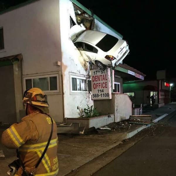 car fail: A white sedan wedged into the second-story wall of a building above a dental office sign with a firefighter watching