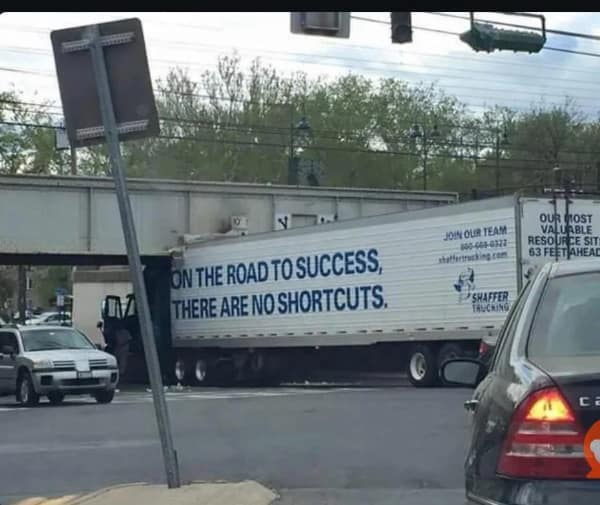 A semi-truck wedged under a low bridge. Text on trailer: "ON THE ROAD TO SUCCESS, THERE ARE NO SHORTCUTS."