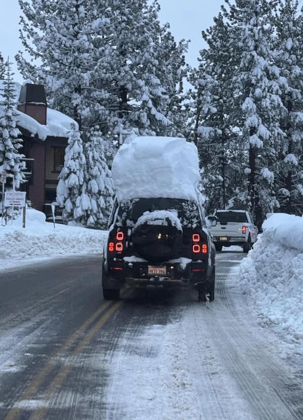 A black SUV driving on a snowy road with a massive, three-foot-thick block of uncleared snow remaining on its roof.